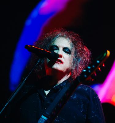 Robert Smith of The Cure with curly hair and dramatic makeup stands on stage, holding a microphone close. The background features colorful, blurred lights, creating a vibrant atmosphere. The person is wearing dark clothing.
