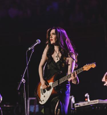 A young woman with long dark hair stands on stage with a guitar