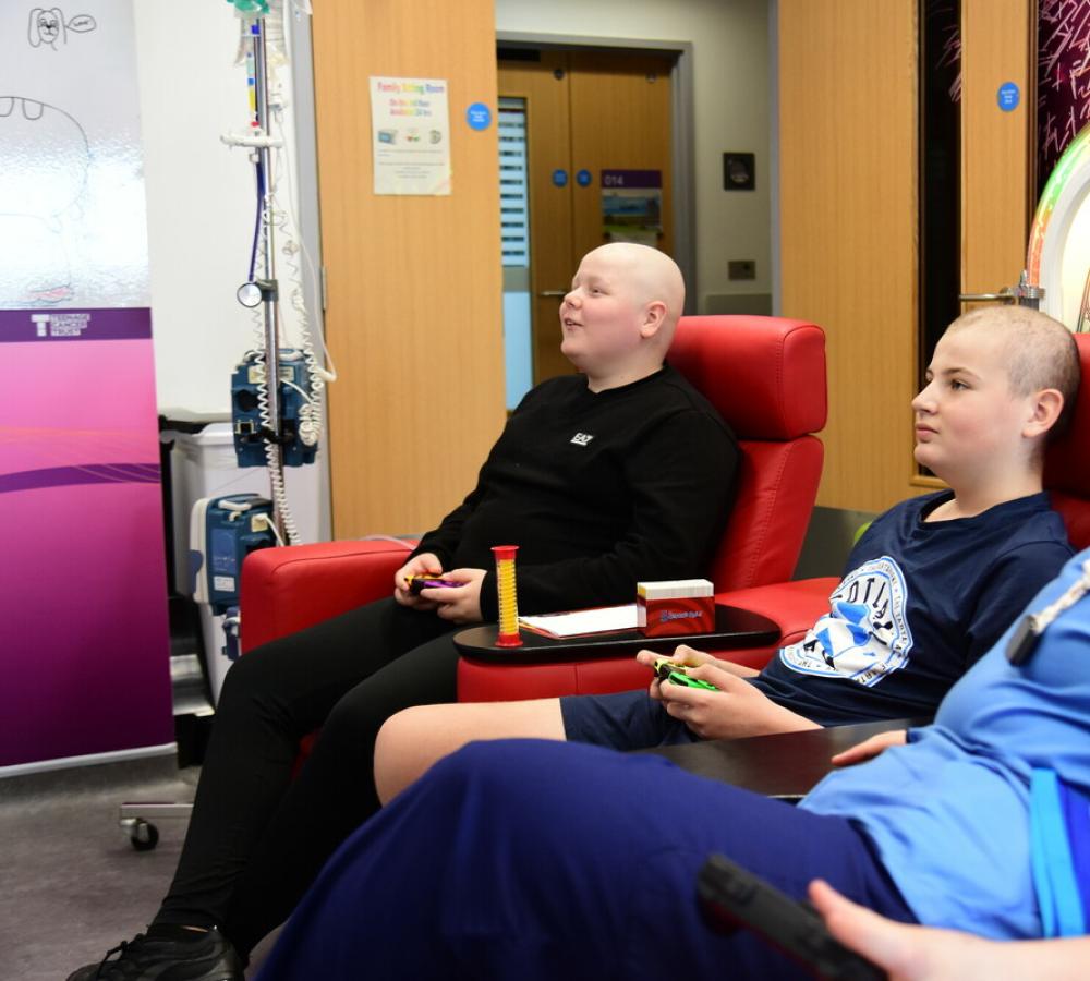 Two young people playing videogames on a Teenage Cancer Trust unit with a nurse watching them play
