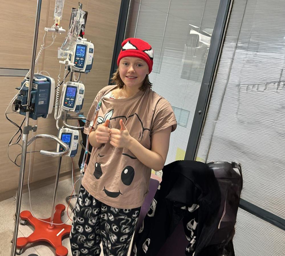 Young person with cancer, attached to a medical drip, undergoing treatment. They are in their pyjamas, wearing a red bandana on their head.