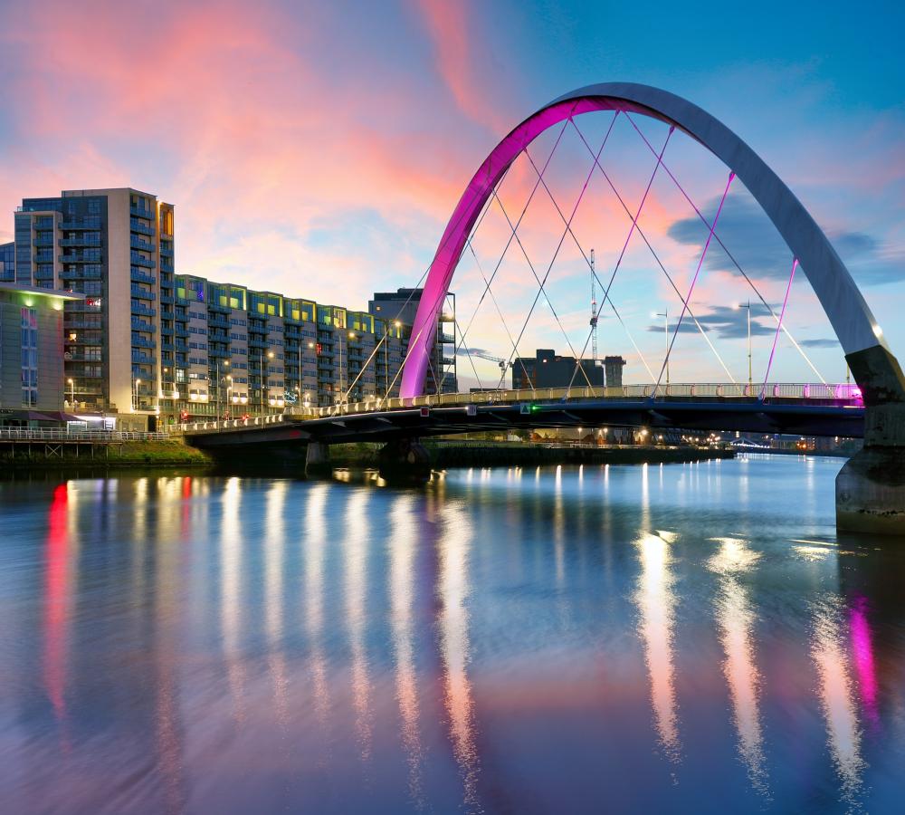 A modern arched bridge with purple lighting spans a calm river at sunset, reflecting city buildings and a colorful sky in the water.