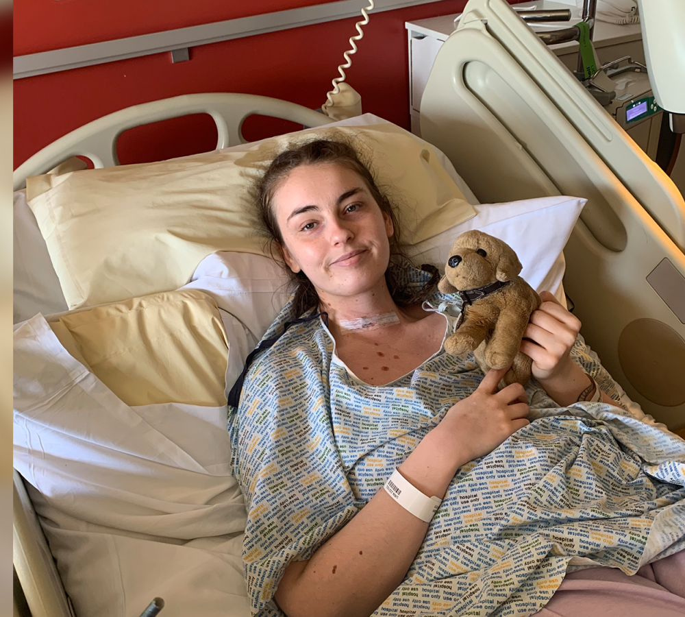 Young woman in a hospital bed smiles at the camera, wearing a hospital gown and holding a small stuffed animal. Medical equipment and pillows are visible around her.