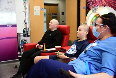 Two young people playing videogames on a Teenage Cancer Trust unit with a nurse watching them play