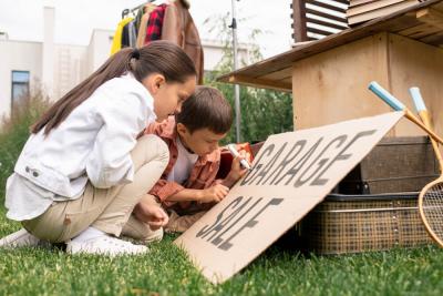 A young girl and a young boy writing the message 'garage sale' on a sign