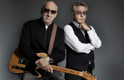 Two men stand side by side against a dark background, embodying the spirit of legendary gigs at Royal Albert Hall. One man (Pete Townsend), in sunglasses and a black jacket, holds a guitar. The other (Roger Daltry), also in shades, wears a white shirt with a black vest and thoughtfully rests his hand on his chin.