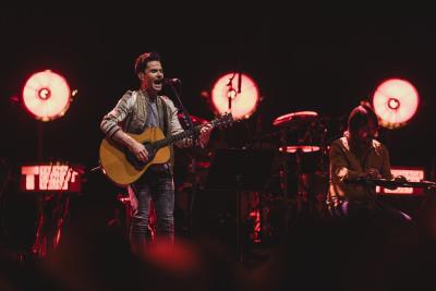 Musician Kelly Jones on stags. He is wearing a grey jacket and playing an acoustic guitar. There are three large spotlights behind him. 