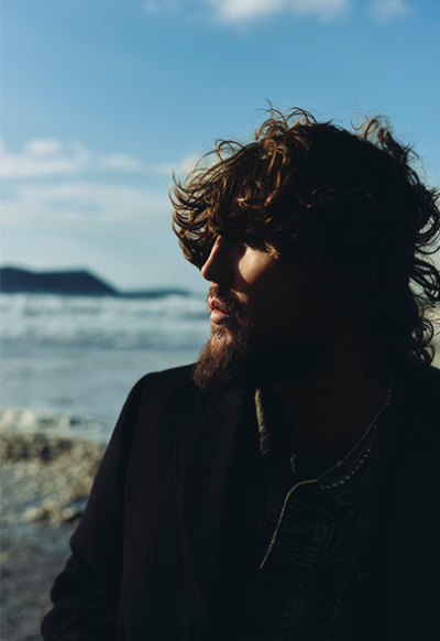 James Arthur with curly hair and a beard stands on a beach, facing sideways. The background features a calm sea and distant hills under a lightly clouded sky. As if caught in the anticipation of 2024 gigs at the Royal Albert Hall, sunlight casts shadows, emphasizing the persons profile.