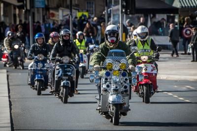 Seven people riding scooters down a road together in the sunshine