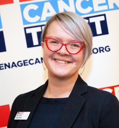 A woman with short blonde hair and red cateye shaped glasses wearing a black top and black jacket
