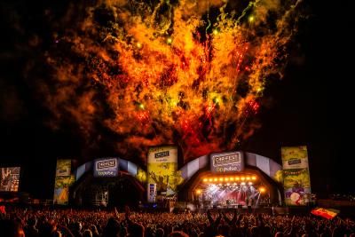 A large crowd watches a band perform on the main stage at CarFest in front of a fireworks display