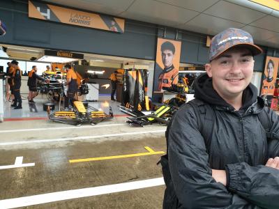 Adam Stuart, Teenage Cancer Trust CarFest ambassador, stands in front of Lando Norris's Formula 1 car, arms folded and smiling