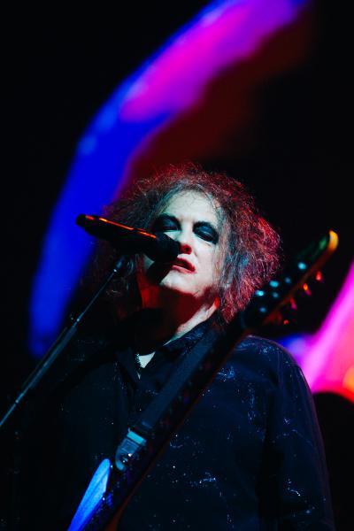 Robert Smith of The Cure with curly hair and dramatic makeup stands on stage, holding a microphone close. The background features colorful, blurred lights, creating a vibrant atmosphere. The person is wearing dark clothing.
