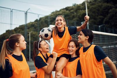 A five-a-side football team wearing orange bibs holds a player on their shoulders, she is holding a ball and celebrating