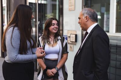 Two young women, both with dark hair, speak animatedly to a male MP wearing a dark suit. He looks like he is asking the women a question and they are replying. 
