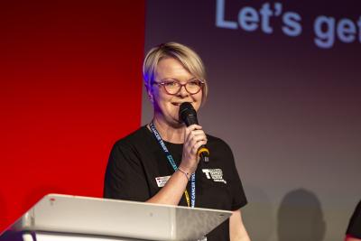 Woman with blonde hair, red framed glasses and a black t-shirt holds a microphone on stage