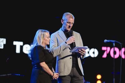 A couple stand on stage speaking into a microphone. The woman has blonde hair and is wearing a black dress, the man is also blonde, he is wearing a light grey jacket