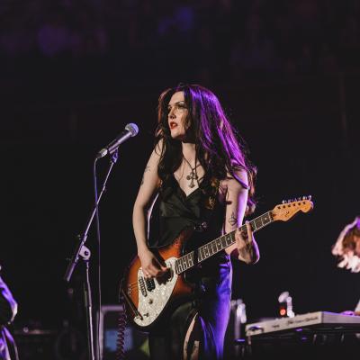 A young woman with long dark hair stands on stage with a guitar