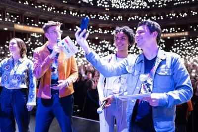 A young woman with curly dark hair takes a selfie with two young men with light brown hair in front of the Royal Albert Hall audience. Phone torches are being used to make a sparkling background