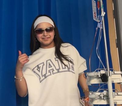 A young woman with brown hair wearing sunglasses gives a thumbs up sign on a hospital unit