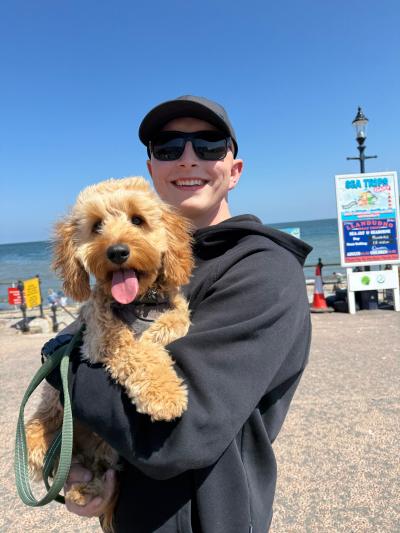 A young man wearing a baseball cap and sunglasses stands smiling on a beach. He is holding a fluffy golden dog, who is also smiling. 