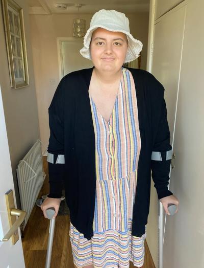 Abbie stands using crutches and wearing a hat at home during cancer treatment