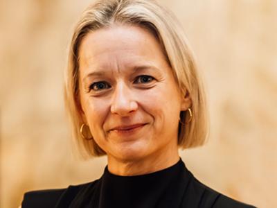 Woman with neck-length blonde hair and black blouse looking at camera with a light brown backdrop behind her.