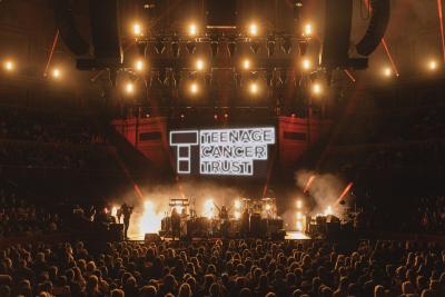 Large hall with crowd flooded with lights and sign on stage reading 'Teenage Cancer Trust'