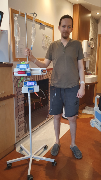 Man in khaki top and grey shorts holding large medical device, including a drip.