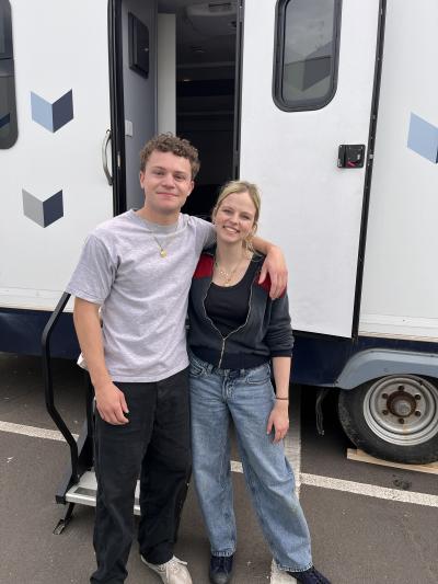 A brown haired young man and a blonde young woman stand outside a trailer on a film set