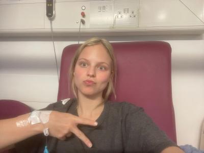 A young woman with blonde hair sits in a hospital chair, she has a cannula in her hand