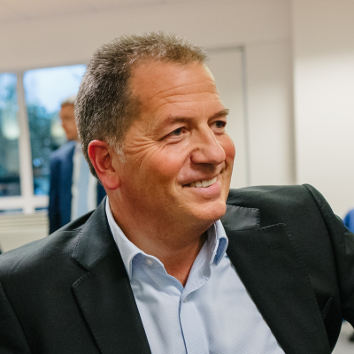 A man in a suit smiles while sitting indoors, with another person blurred in the background and windows letting in natural light.