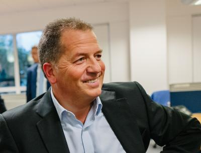 A man in a suit smiles while sitting in an office setting, with desks and windows visible in the background.