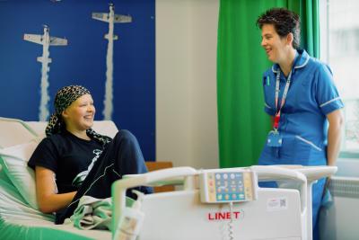 Young woman wearing headscarf sat on hospital bed smiling and chatting to nurse on Teenage Cancer Trust unit